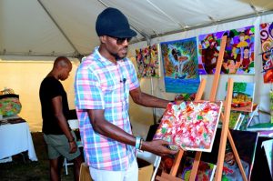 Artist Ralston Vassell pauses to show an abstract piece in progress during live art demo, as a patron browses his exhibit.